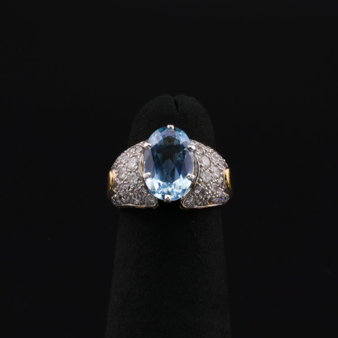 A close-up of a two-toned ring with platinum and yellow gold chunky pave ring featuring a stunning large aquamarine center stone with an array of smaller oval diamonds resting on a black ring holder against a black background.