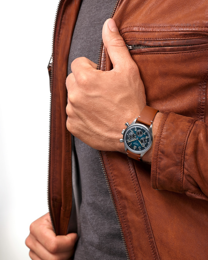 A close-up of a man holding his hand up to his leather jacket with a watch around his wrist. The watch features a blue & black dial, tan & white hands and markers, a stainless steel bezel, three crowns, and a brown leather strap.