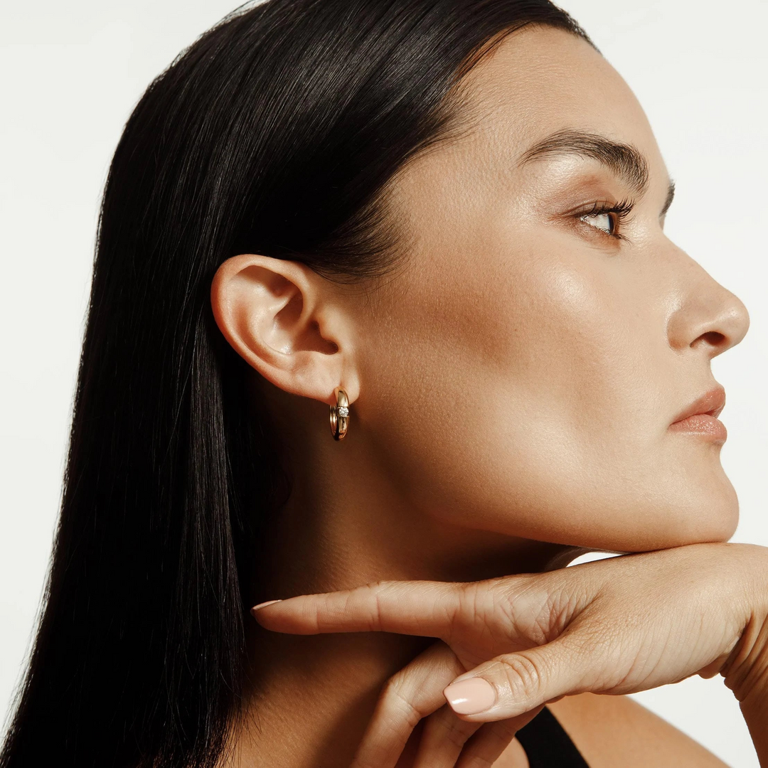 A close-up of a woman wearing a yellow-gold earring. The rounded hoop earrings feature a floating diamond in the center.