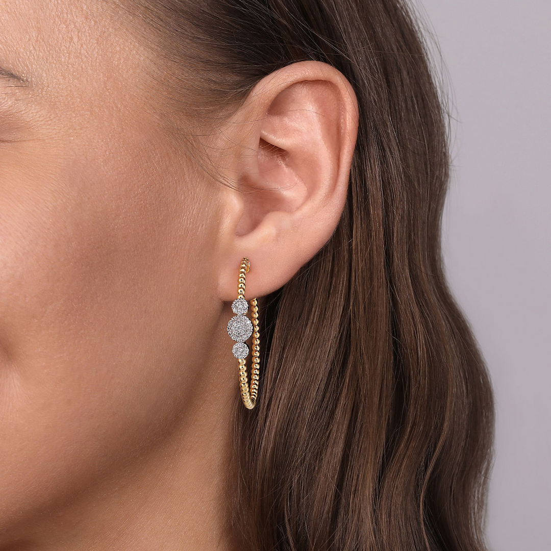 A close-up of a woman with brunette hair wearing diamond hoop earrings with circles of golden beads are crowned with a trio of diamond circles