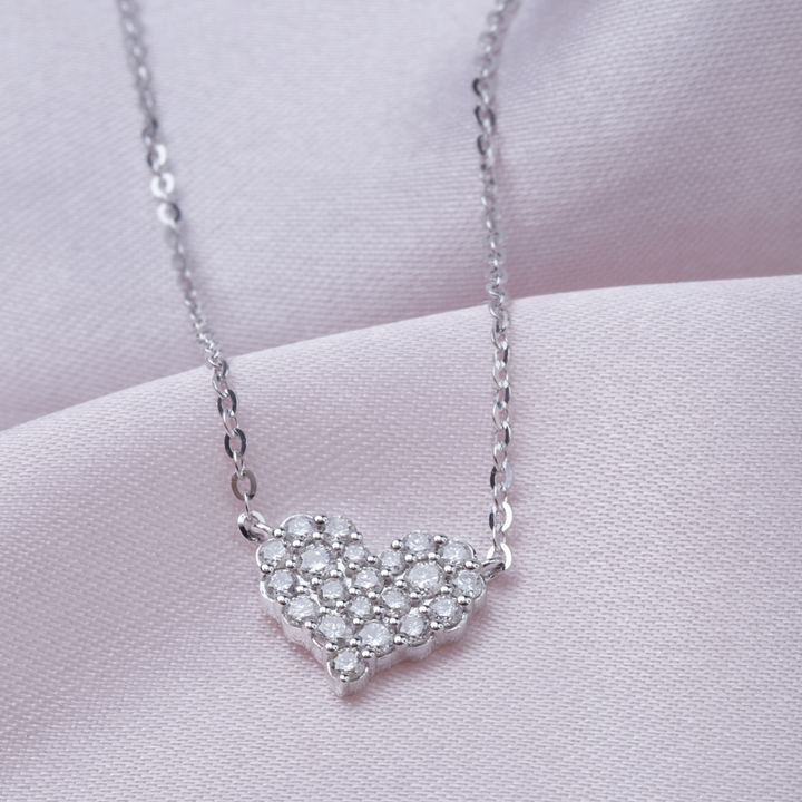 A close-up of a white-gold Diamond Cluster Heart Necklace against a purple background. The necklace features a heart design with a cluster of round diamonds.