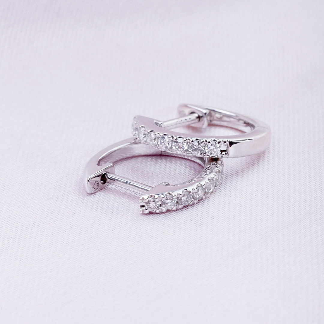 A pair of U-shaped huggies with a single row of round diamonds rests against a light pink background.