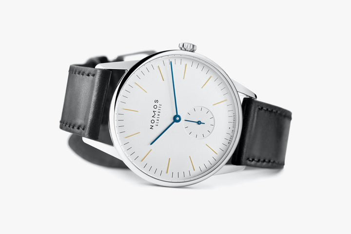 A Nomos Glashutte watch resting on the left side against a white background. It features a white dial, gold & blue hands and markers, a stainless steel bezel, and a black strap.