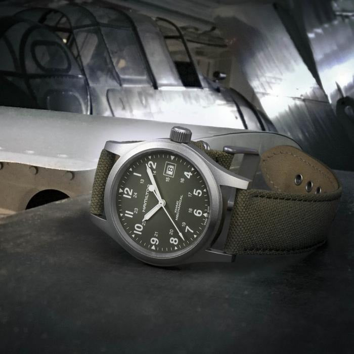 A Hamilton watch is lying on its left side next to a plane. It features a green dial, white hands and markers, a stainless steel case, a steel bezel, a steel crown, and a green canvas strap.