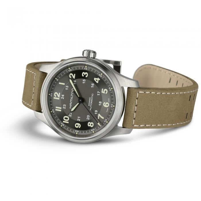 A Hamilton watch is lying on its left side against a white background. It features a black dial, light green hands and markers, a stainless steel case, bezel, and crown, and a brown calf leather strap.