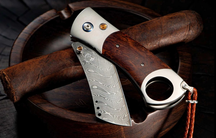 A close-up of a pocket knife with the blade out. The pocket knife features a titanium Cigar Cutter with a Desert Ironwood handle and a Damascus steel blade.