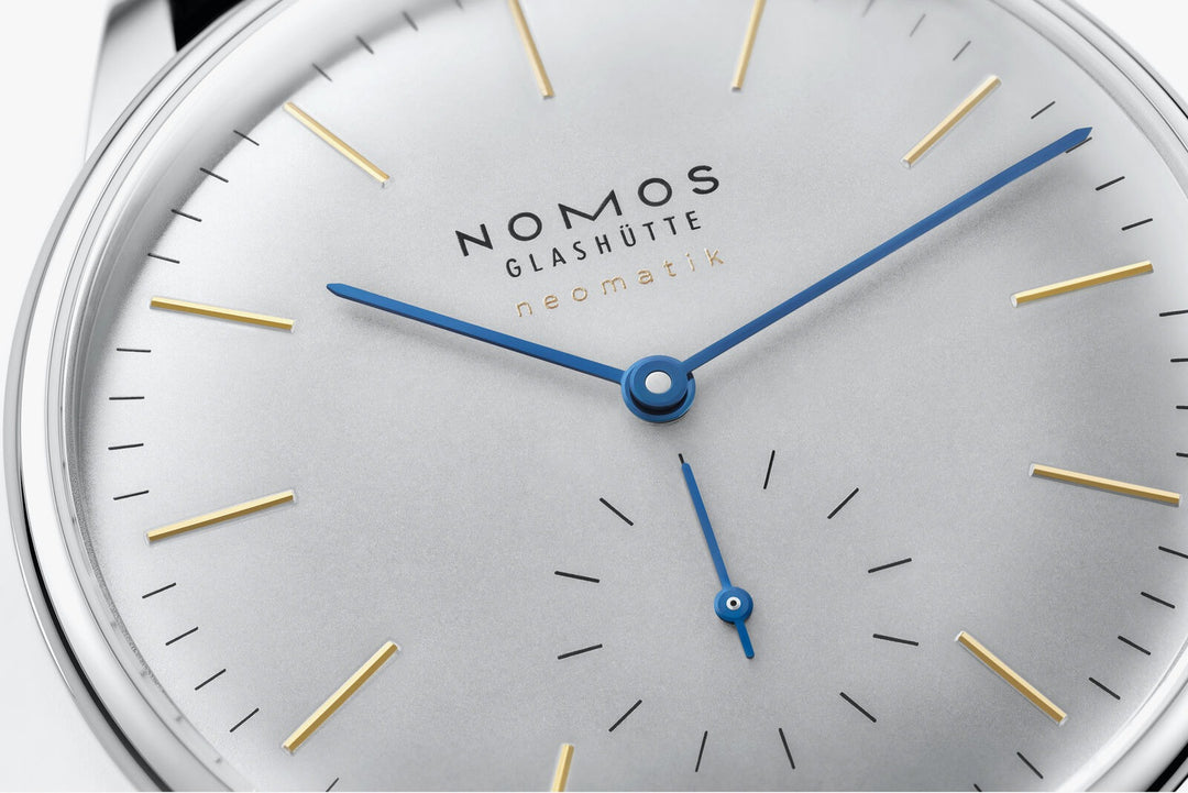 A close-up image of a Nomos Glashutte watch angled to the left, focusing on the dial. It features a white dial, gold & blue hands, and markers, and a stainless steel bezel.