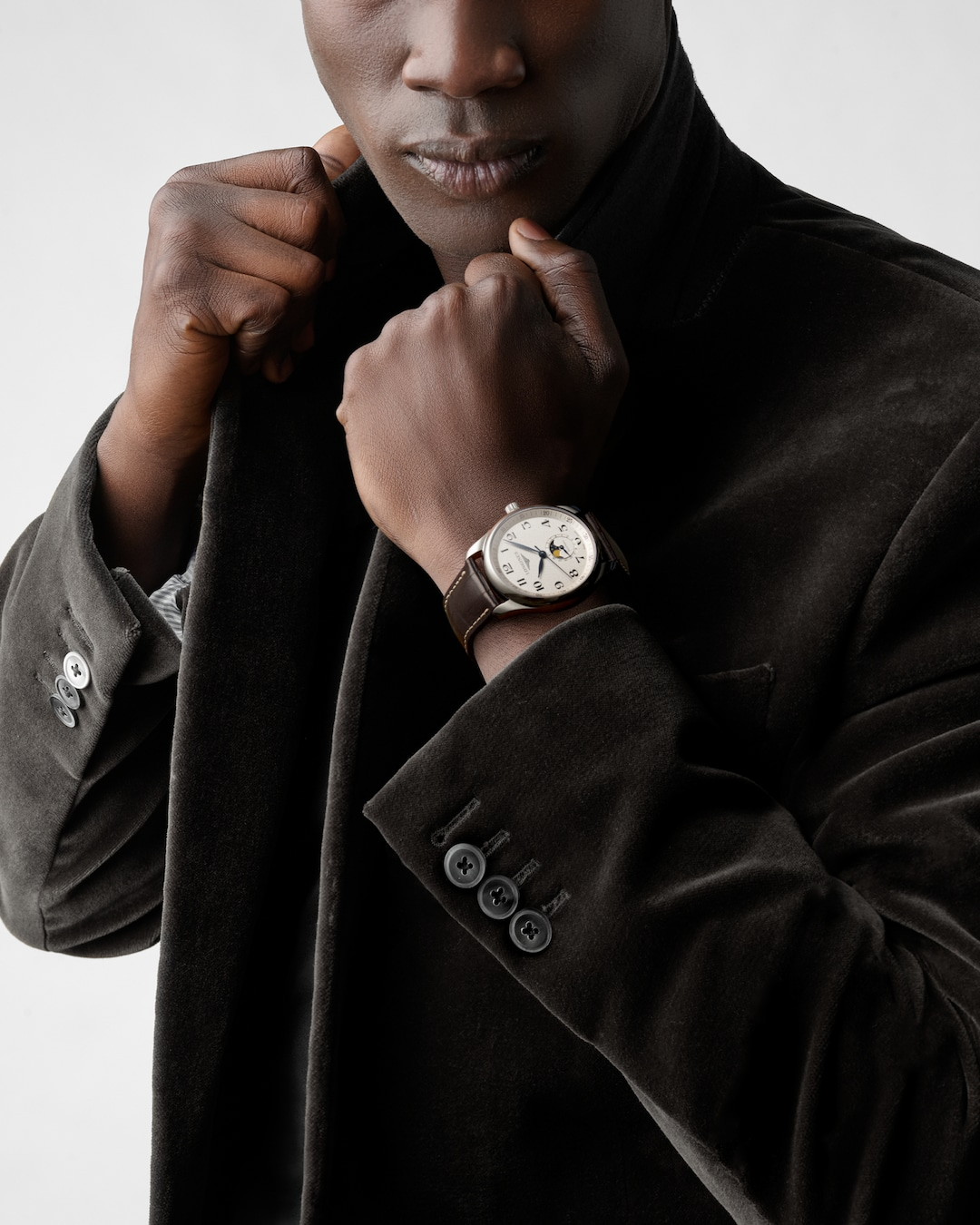 A close-up of a man holding his hand to his jacket collar with a watch around his wrist. The watch features a silver dial, black & blue hands and markers, a stainless steel bezel, a crown on the right side, and a brown leather strap.