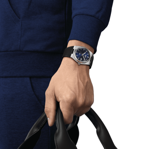 A man wearing a Tissot watch around his wrist. The watch features a navy dial, a steel bezel, white hands and markers, and a black strap.