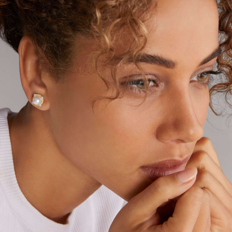 A woman is wearing 18K gold and sterling silver stud earrings with a freshwater cultured pearl accented by diamonds and caviar beading