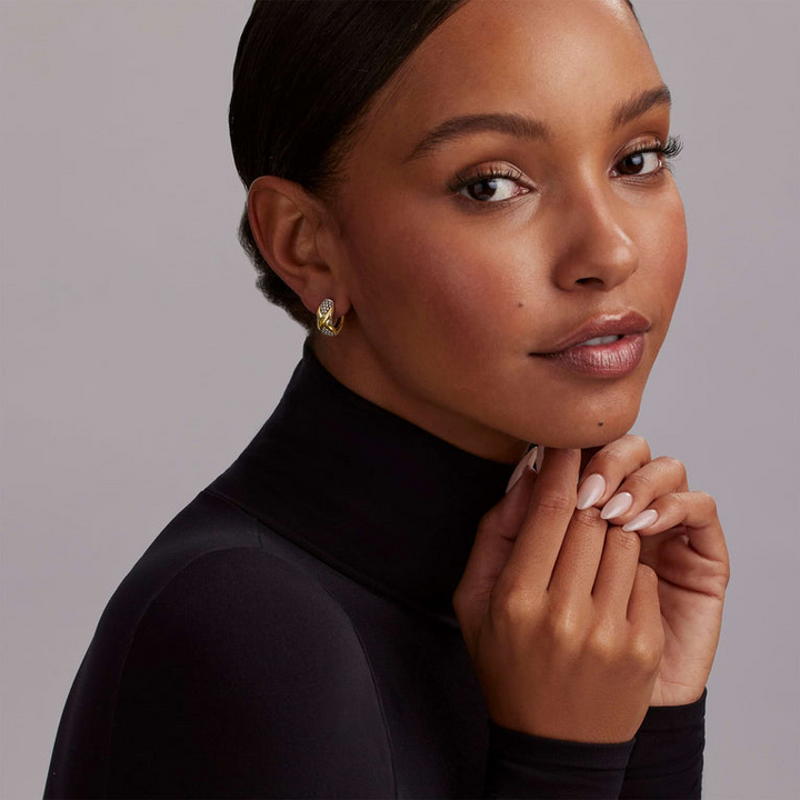 A woman is wearing a black turtleneck and a pair of two-tone huggie earrings with Smooth 18k gold and sterling silver Caviar beading and an x motif.