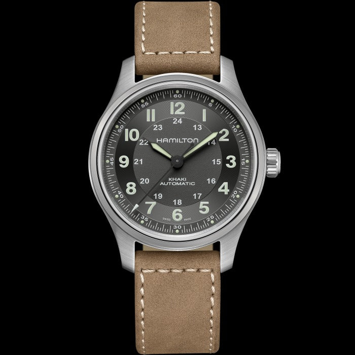 A Hamilton watch is laid out against a black background. It features a black dial, light green hands and markers, a stainless steel case, bezel, and crown, and a brown calf leather strap.