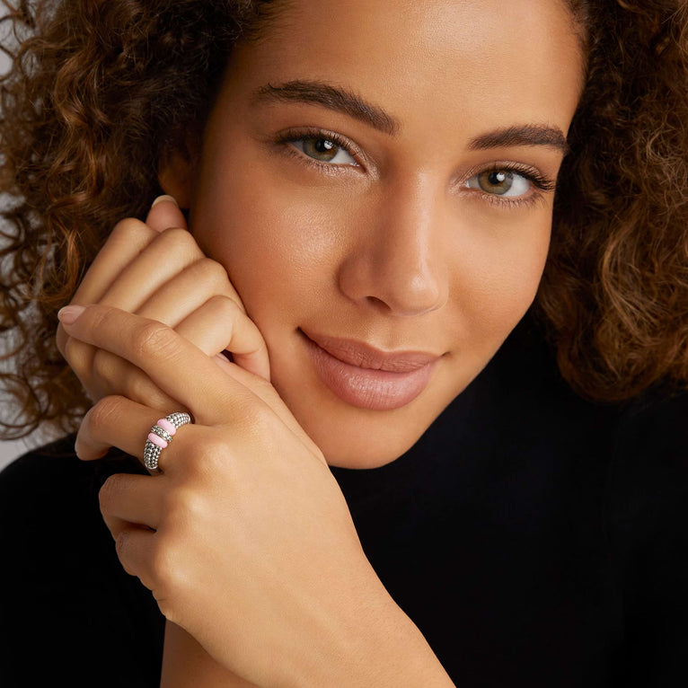 A close-up of a woman with brown hair holding her hands up to her cheek showcasing the ring on her middle finger. The ring features caviar beading, two light pink bar stations, and a diamond-set bar station in the middle.