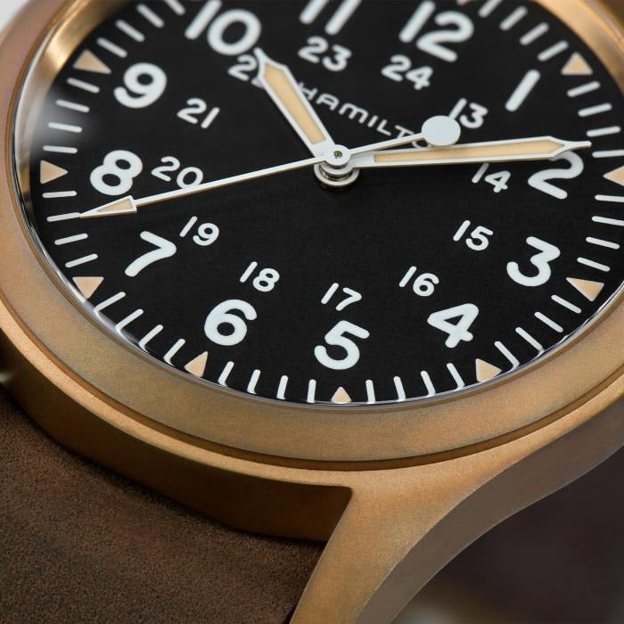 A close-up of a Hamilton watch, focusing on the dial, bronze case, a bronze crown, and a brown leather strap.