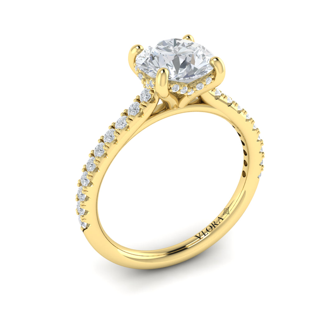 An angled view of a yellow gold engagement ring is set against a white background. The engagement ring features a round center diamond, a hidden halo, and a diamond-set band.