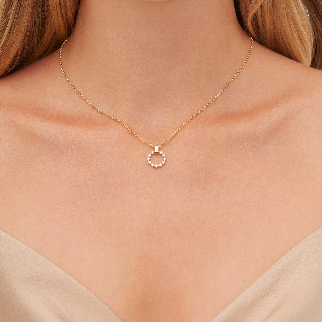 A close-up of a woman wearing a beige blouse and a yellow necklace with a diamond-set ring pendant. The ring features a shared prong motif and is suspended from a smooth gold bar bail.