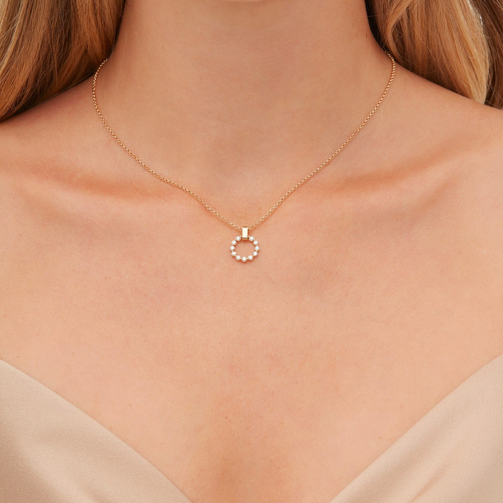 A close-up of a woman wearing a beige blouse and a yellow necklace with a diamond-set ring pendant. The ring features a shared prong motif and is suspended from a smooth gold bar bail.