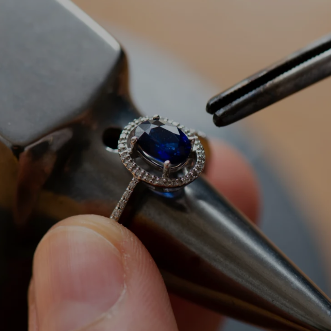 A close-up of a ring being held in a jeweler's loupe and tweezers. The ring features a round deep blue gemstone surrounded by a halo of small clear stones, set on a silver-toned band.