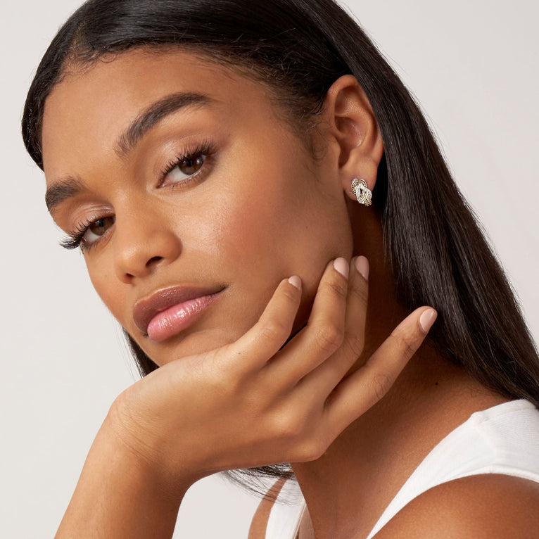 A woman is wearing a white tank top and a pair of omega clip huggie earrings with diamonds and 18K gold woven in a knot motif. 