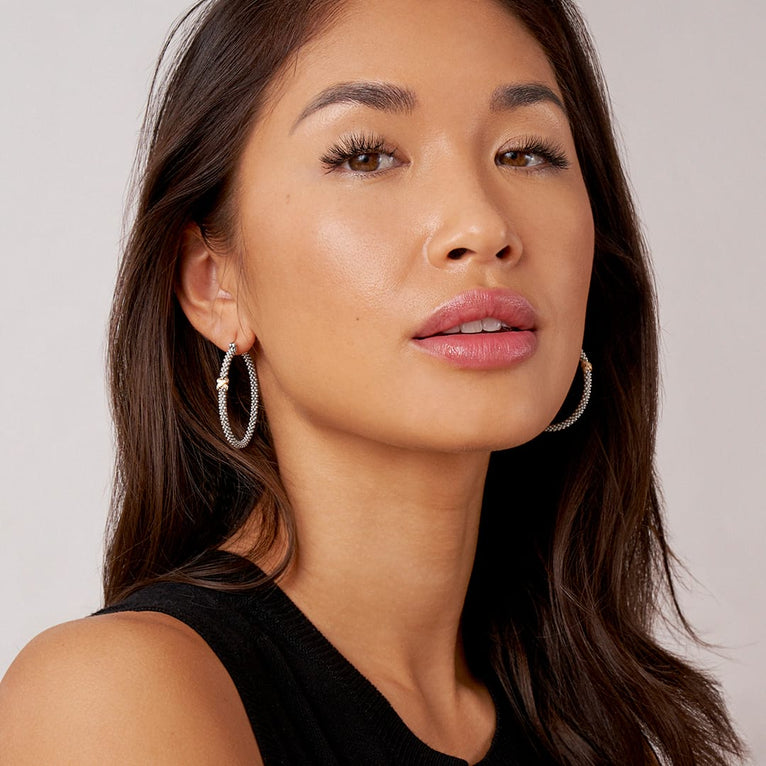 A woman is wearing a black top and a pair of hoop earrings with a diamond and 18K gold x station with sterling silver caviar beading. 