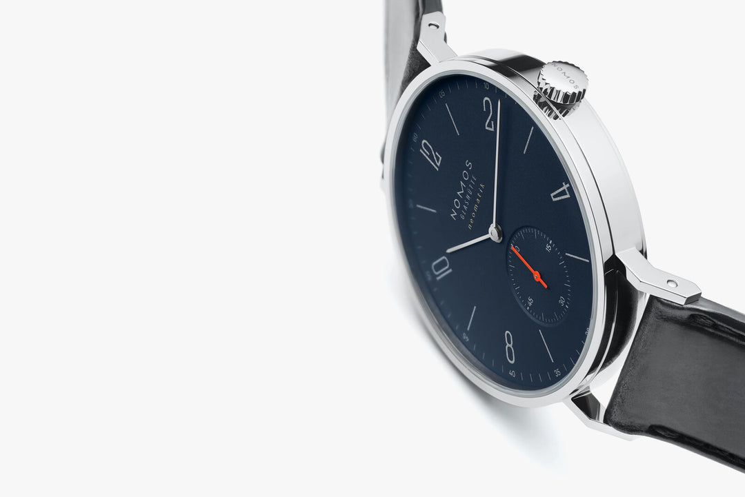 A Nomos Glashutte watch resting on the left side against a white background. It features a dark blue dial, white hands and markers, a stainless steel bezel, and a black strap.