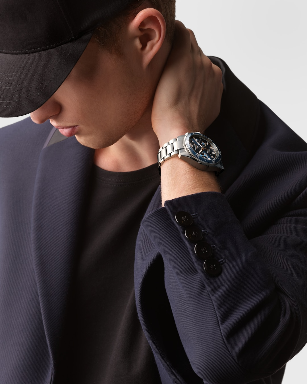 A close-up of a man holding his hand to his beck with a watch around his wrist. The watch features a black & blue dial, white & gold hands and markers, a blue bezel, three crowns on the right side, and a stainless steel bracelet.