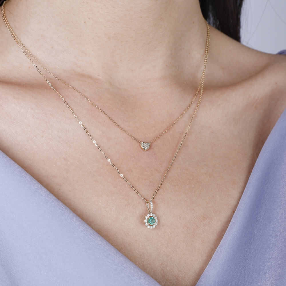 A woman is wearing a yellow-gold Halo Necklace and a purple top. The necklace features an emerald round-cut center stone surrounded by round diamonds. 