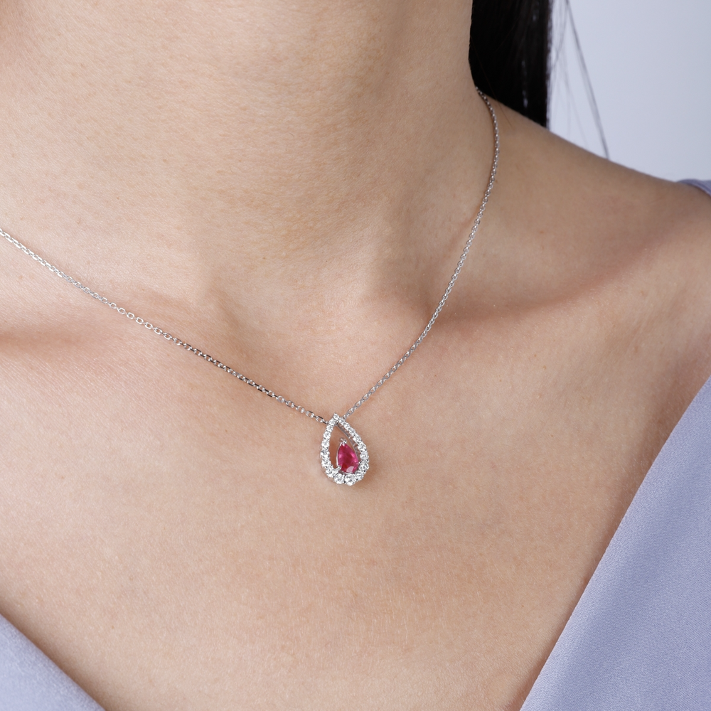 A woman is wearing a  white-gold Diamond Teardrop Necklace and a purple top. The necklace features a ruby teardrop-shaped diamond design with a halo of round diamonds.