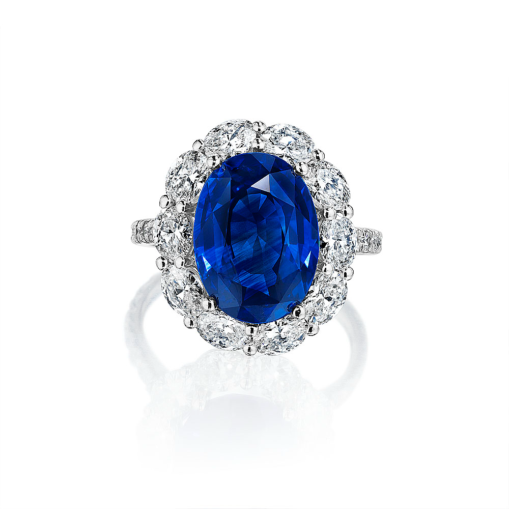 A front view of a white gold diamond ring against a white background. The ring features a Sri Lankan vivid blue oval-cut sapphire center stone, framed by a halo made of oval-cut diamonds, and accented by a row of round brilliant-cut diamonds set along the shank.