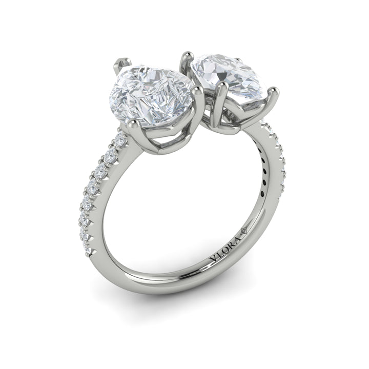 An angled view of a white gold engagement ring is set against a white background. The engagement ring features an oval and pear-shaped diamond meeting in the center, surrounded by a half-eternity diamond band.