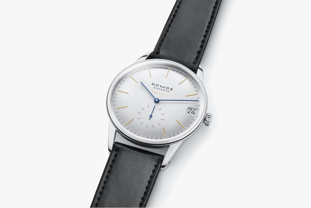 A close-up image of a Nomos Glashutte watch angled to the right. It features a white dial, gold & blue hands and markers, a stainless steel bezel, and a black strap.