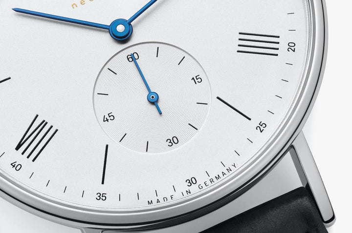 A close-up image of a Nomos Glashutte watch angled to the left, focusing on the dial. It features a white dial, blue hands and markers, a stainless steel bezel, and a black strap.