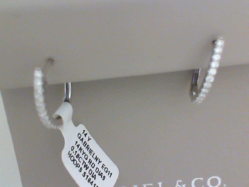 A close-up of two paves of diamond huggies rests on a Gabriel & Co gray display with a tag around the left earring.