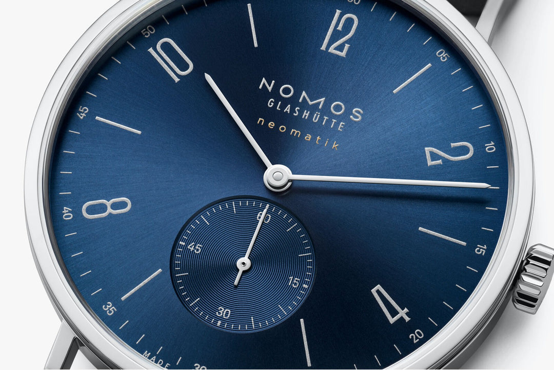 This is a close-up image of a Nomos Glashutte watch angled to the right, focusing on the dial. It features a blue dial, silver hands and markers, a stainless steel bezel, and a black leather strap.