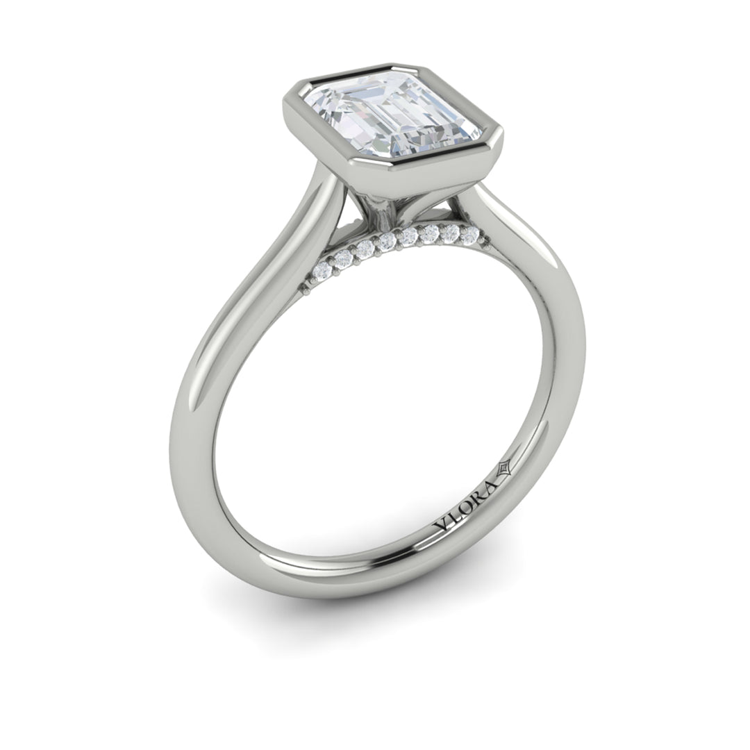 An angled view of a white gold engagement ring is set against a white background. The engagement ring features a sleek bezel that frames the emerald cut diamond, rising in a cathedral-style setting and Diamond pavé accents beneath the center stone.