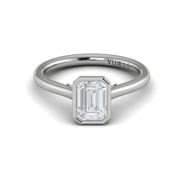 A front view of a white gold engagement ring is set against a white background. The engagement ring features a sleek bezel that frames the emerald cut diamond, rising in a cathedral-style setting and Diamond pavé accents beneath the center stone.