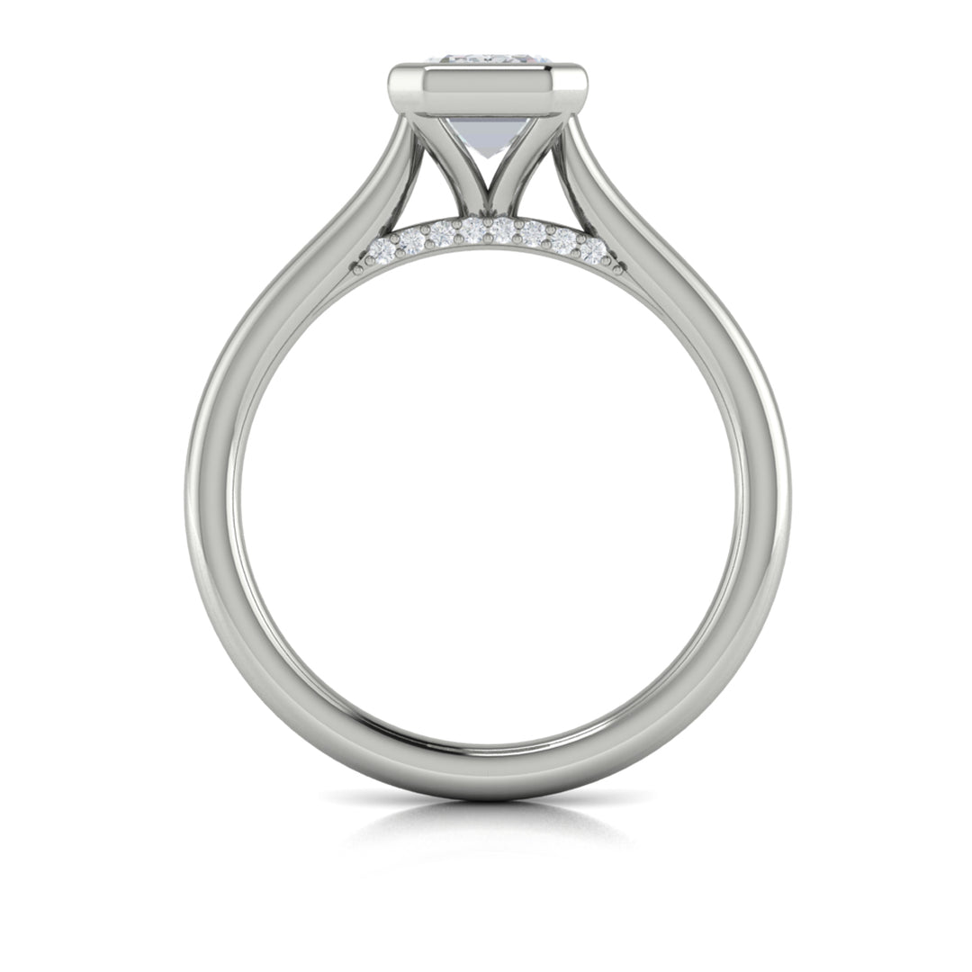A top view of a white gold engagement ring is set against a white background. The engagement ring features a sleek bezel that frames the emerald cut diamond, rising in a cathedral-style setting and Diamond pavé accents beneath the center stone.
