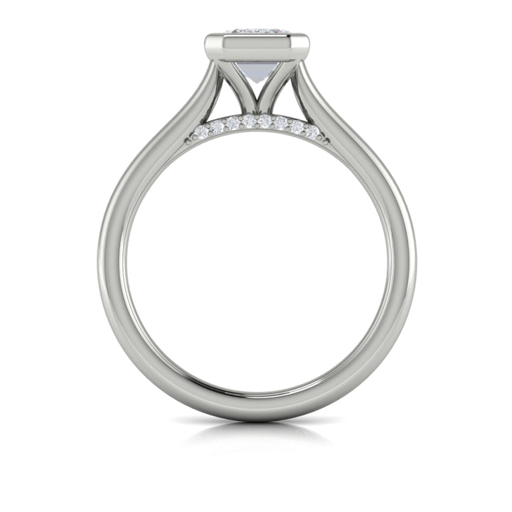 A top view of a white gold engagement ring is set against a white background. The engagement ring features a sleek bezel that frames the emerald cut diamond, rising in a cathedral-style setting and Diamond pavé accents beneath the center stone.