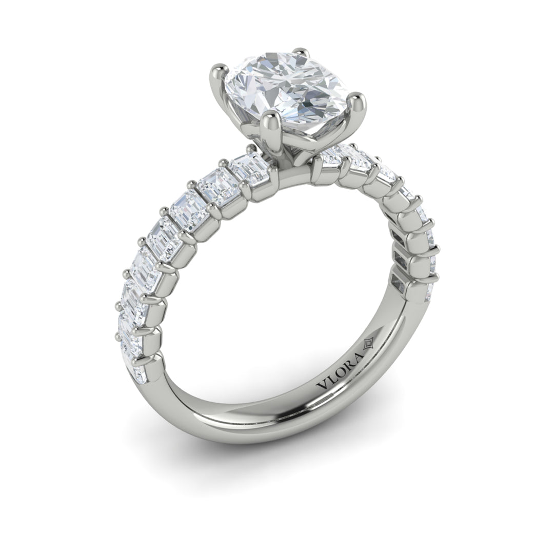 An angled view of a white gold engagement ring is set against a white background. The engagement ring features an oval-shaped center stone, raised in the Vlora signature V-setting, with emerald-cut accent diamonds blazing across the band.