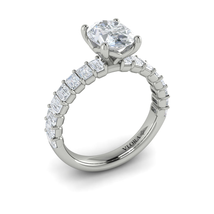 An angled view of a white gold engagement ring is set against a white background. The engagement ring features an oval-shaped center stone, raised in the Vlora signature V-setting, with emerald-cut accent diamonds blazing across the band.