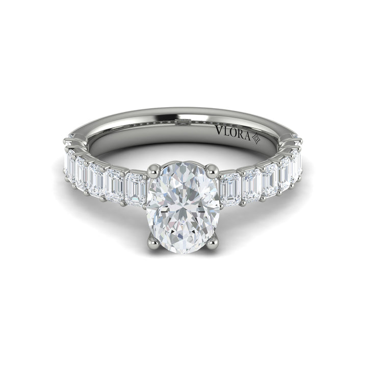A front view of a white gold engagement ring is set against a white background. The engagement ring features an oval-shaped center stone, raised in the Vlora signature V-setting, with emerald-cut accent diamonds blazing across the band.