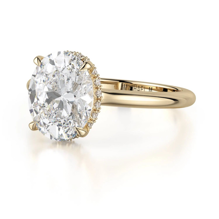 A close-up view of the Hidden Halo Solitaire Engagement Ring, made of yellow gold. The ring is angled against a white background to the left. It features an oval-cut diamond set in the center with a diamond set hidden halo. Showing the "Michael M" inscription is visible inside the band.