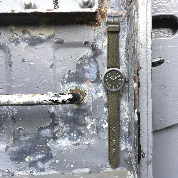 A Hamilton watch is lying on its left side next to a plane. It features a green dial, white hands and markers, a stainless steel case, a steel bezel, a steel crown, and a green canvas strap.