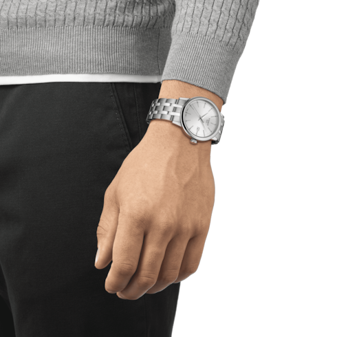 A woman wearing a gray sweater and black pants with a Tissot watch on her wrist. The watch features a silver dial, steel bezel, silver hands and markers, and a stainless steel bracelet.