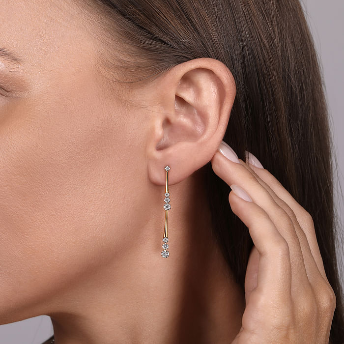 A woman is wearing a pair of drop earrings with 14k yellow gold linear diamond stations and diamond clusters. 