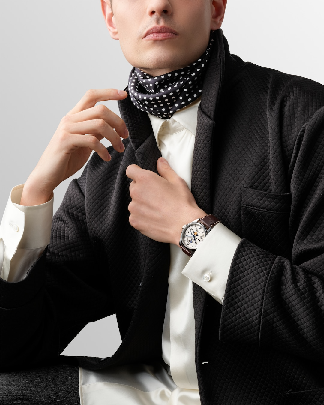 A close-up of a man holding his hand to his jacket with a watch around his wrist. The watch features a silver dial, black & blue hands and markers, a stainless steel bezel, three crowns on the right side, and a brown leather strap.