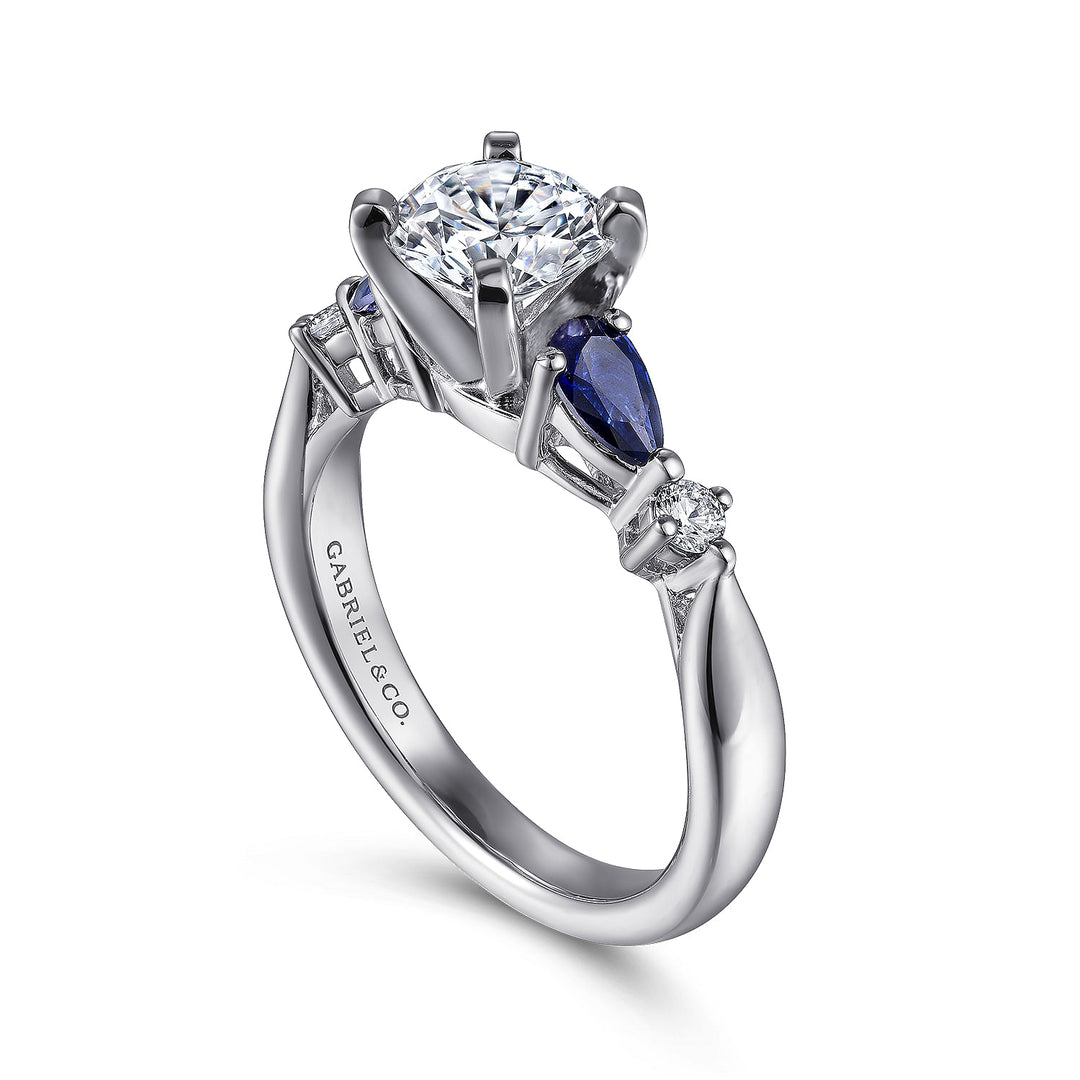 A close-up of a ring made of white gold, angled to the right against a white background. It features a brilliant-cut diamond set in the center with sapphire side stones.