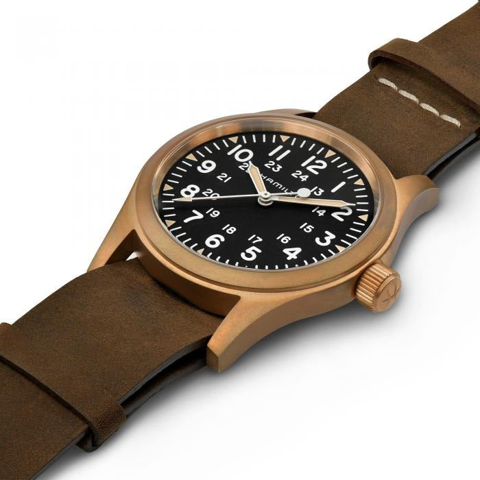 A Hamilton watch is laid out tilted to the right against a white background. It features a black dial, white hands and markers, a bronze case, a bronze bezel, a bronze crown, and a brown nato leather strap.