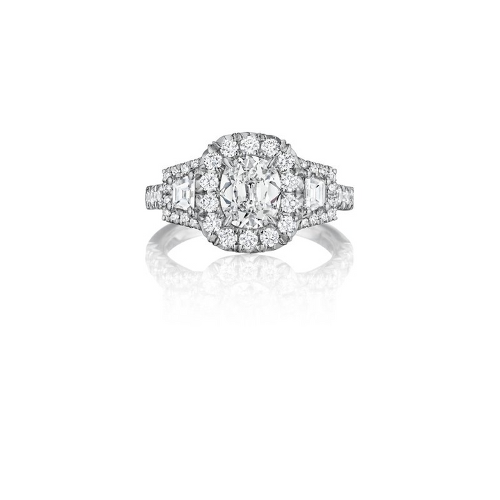 A white gold ring lies flat against a white background. It features a cushion-cut diamond set in the center, halo, and trapezoid side stones.