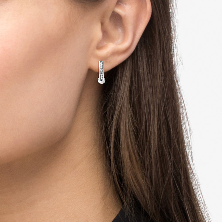 A woman with long brunette hair is wearing a pair of huggie earrings with diamonds set in a smooth sterling silver circle motif.